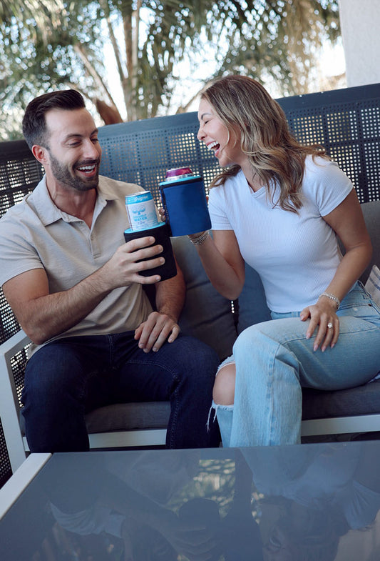 A man and a woman sitting on a patio sofa, laughing and holding iCube Wrapsulator regulars. The man wears a beige polo shirt and dark jeans, while the woman wears a white t-shirt and light-wash ripped jeans.