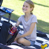 A young girl with blonde hair in a ponytail sits on a black and white picnic blanket at a soccer game on a grassy lawn. She is wearing a gray and white striped t-shirt and denim shorts, smiling as she reaches into a CaddyO Dual Travel Tote containing a bottle of Brisk Fruit Punch