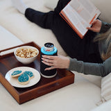 A person lounging on a cream sofa reads a book while holding a black Wrapsulator Slim. A wooden tray on the couch holds a bowl of popcorn, a plate of blue-frosted cookies, and the can cooler.